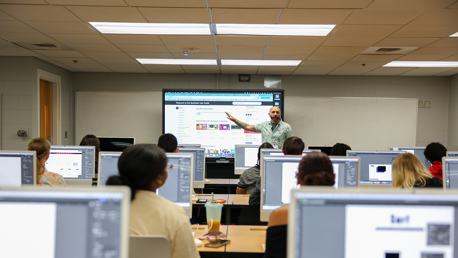 Feature image for the page. Picture of a classroom in progress, with computers at the desks and a professor giving a lesson on a larger screen.