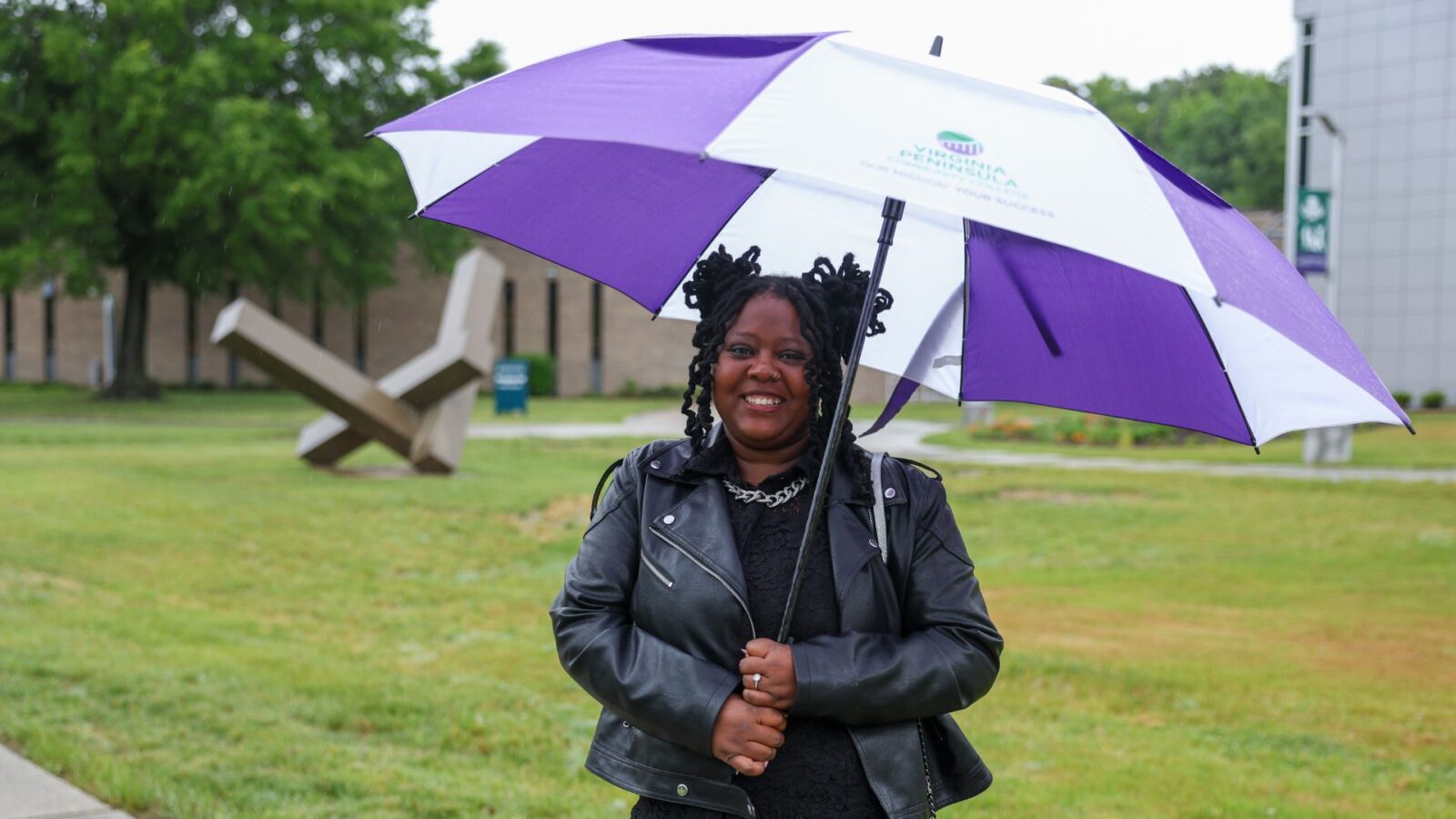 A woman holding an umbrella stands outside with a sculpture in the background.